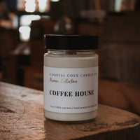 A glass jar candle with a black lid and a white label reading 'COFFEE HOUSE' from Coastal Cove Candle Co., placed on a rustic wooden table in a warm, blurred indoor setting.