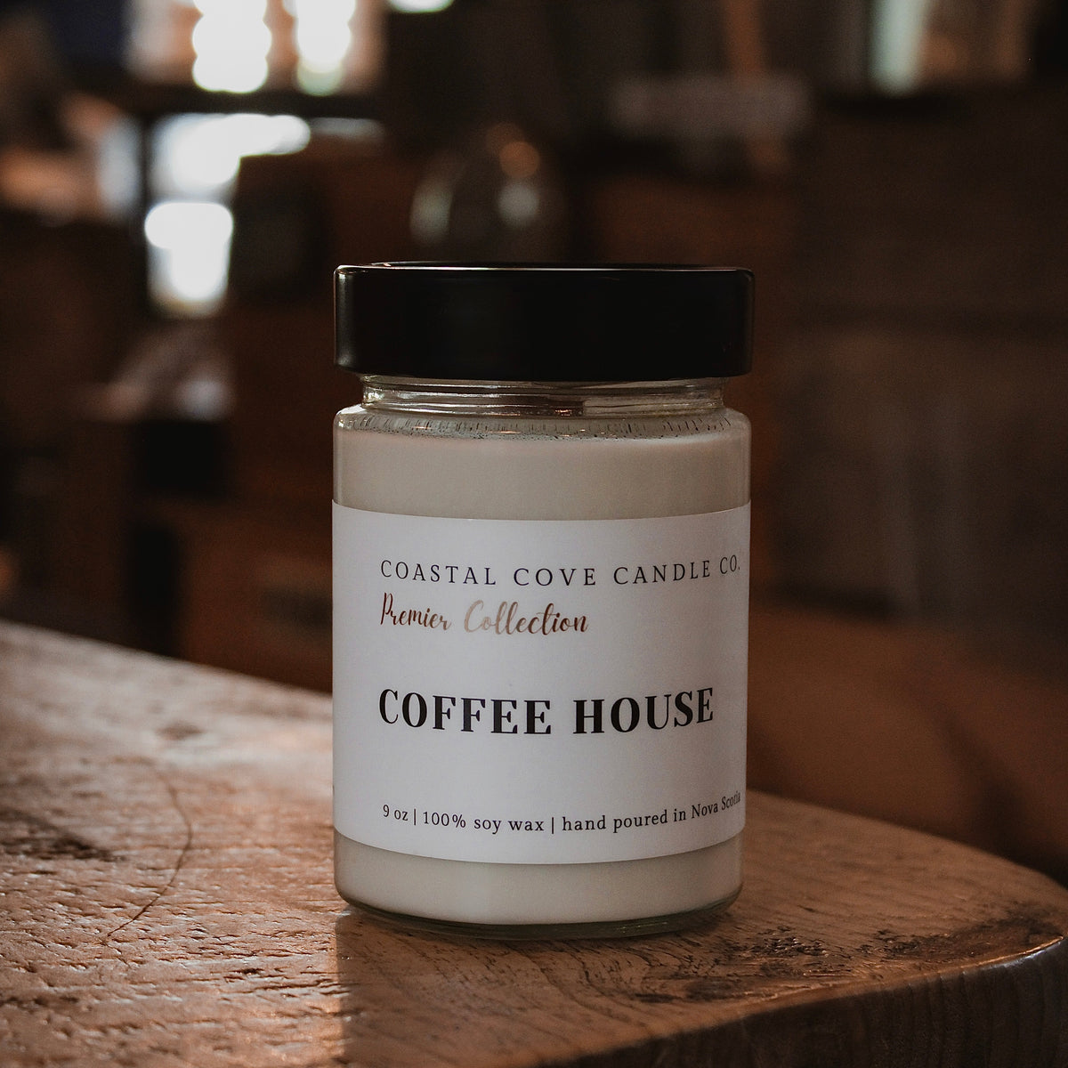 A glass jar candle with a black lid and a white label reading 'COFFEE HOUSE' from Coastal Cove Candle Co., placed on a rustic wooden table in a warm, blurred indoor setting.