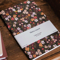 A floral mini notebook with a white paper band label reading 'Mini & Luxe', photographed on a rustic wooden surface.