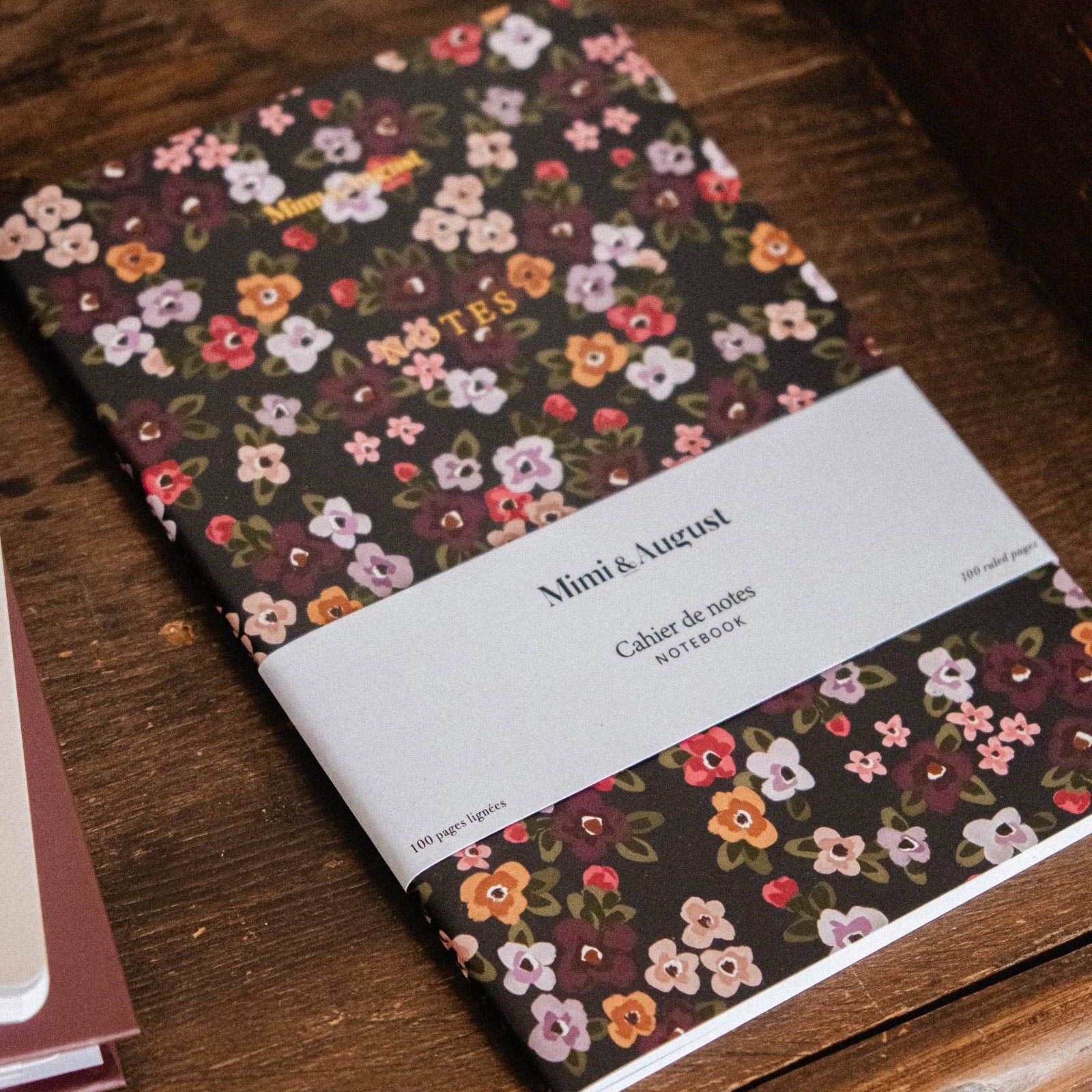A floral mini notebook with a white paper band label reading 'Mini & Luxe', photographed on a rustic wooden surface.