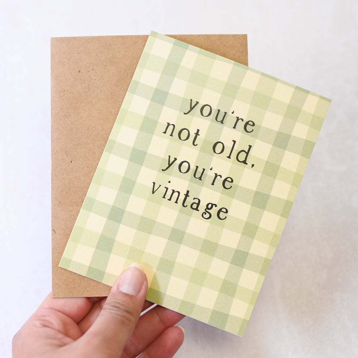 Hand holding a green checkered card with text 'you're not old, you're vintage' on a light background