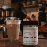 A jar candle labeled 'COFFEE HOUSE' by Coastal Cove Candle Co sits on a wooden counter in a cafe, next to an iced coffee.
