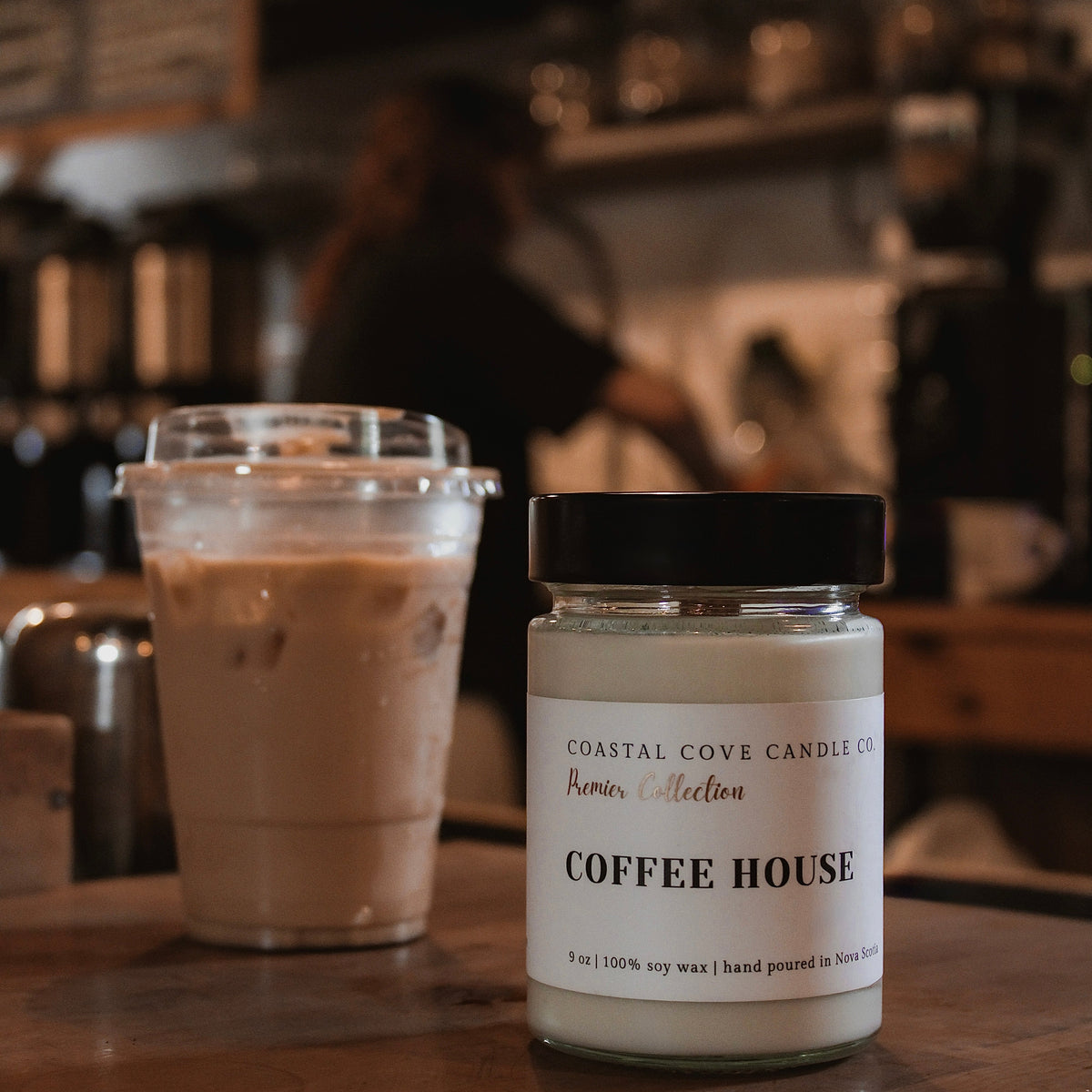A jar candle labeled 'COFFEE HOUSE' by Coastal Cove Candle Co sits on a wooden counter in a cafe, next to an iced coffee.