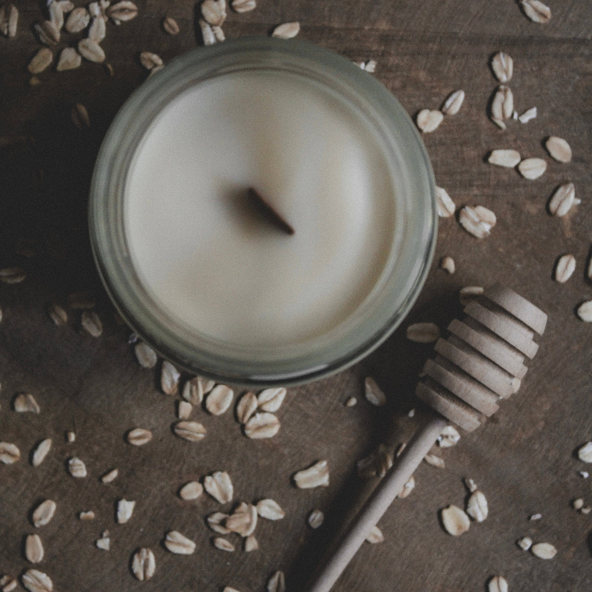 A round glass jar candle with white wax sits on a dark wooden surface scattered with oats, alongside a wooden honey dipper.