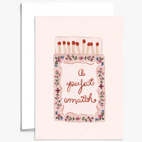 A greeting card with a light pink background featuring an illustration of a matchbox with flowers around the border and the words 'A perfect match' in cursive. A white envelope is partially visible behind the card.