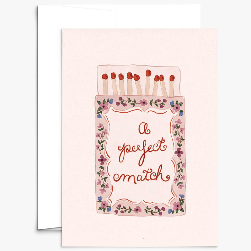 A greeting card with a light pink background featuring an illustration of a matchbox with flowers around the border and the words 'A perfect match' in cursive. A white envelope is partially visible behind the card.