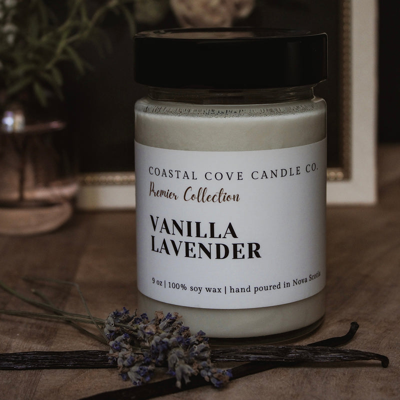 A Coastal Cove Candle Co. Vanilla Lavender soy wax candle in a clear glass jar with a black lid, styled on a wooden surface with dried lavender and vanilla beans in front of a framed floral print.