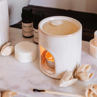 White ceramic diffuser with a lit candle on a marble surface with essential oils and decorative items.