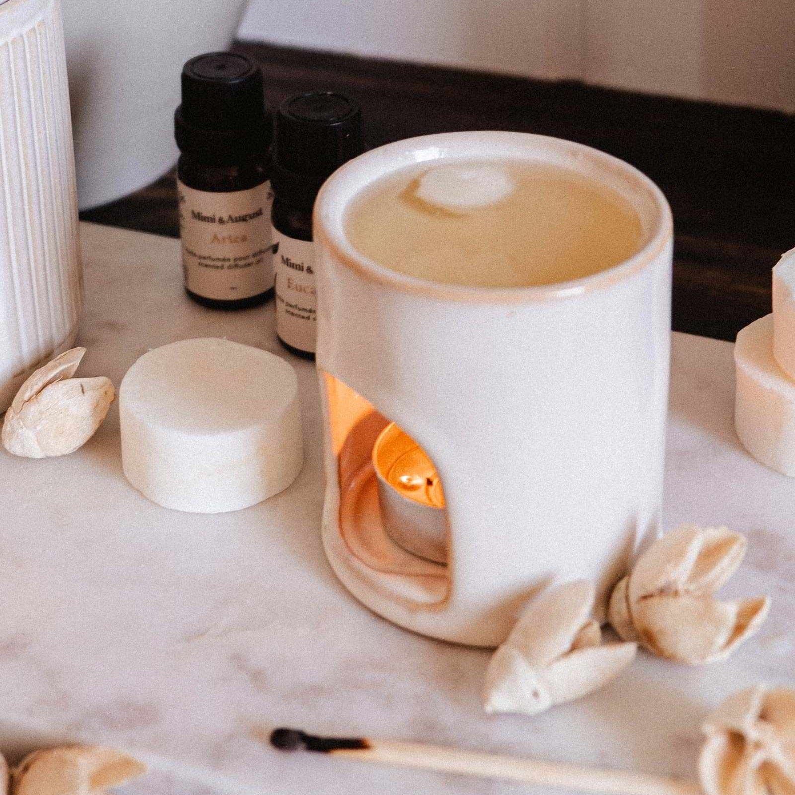 White ceramic diffuser with a lit candle on a marble surface with essential oils and decorative items.