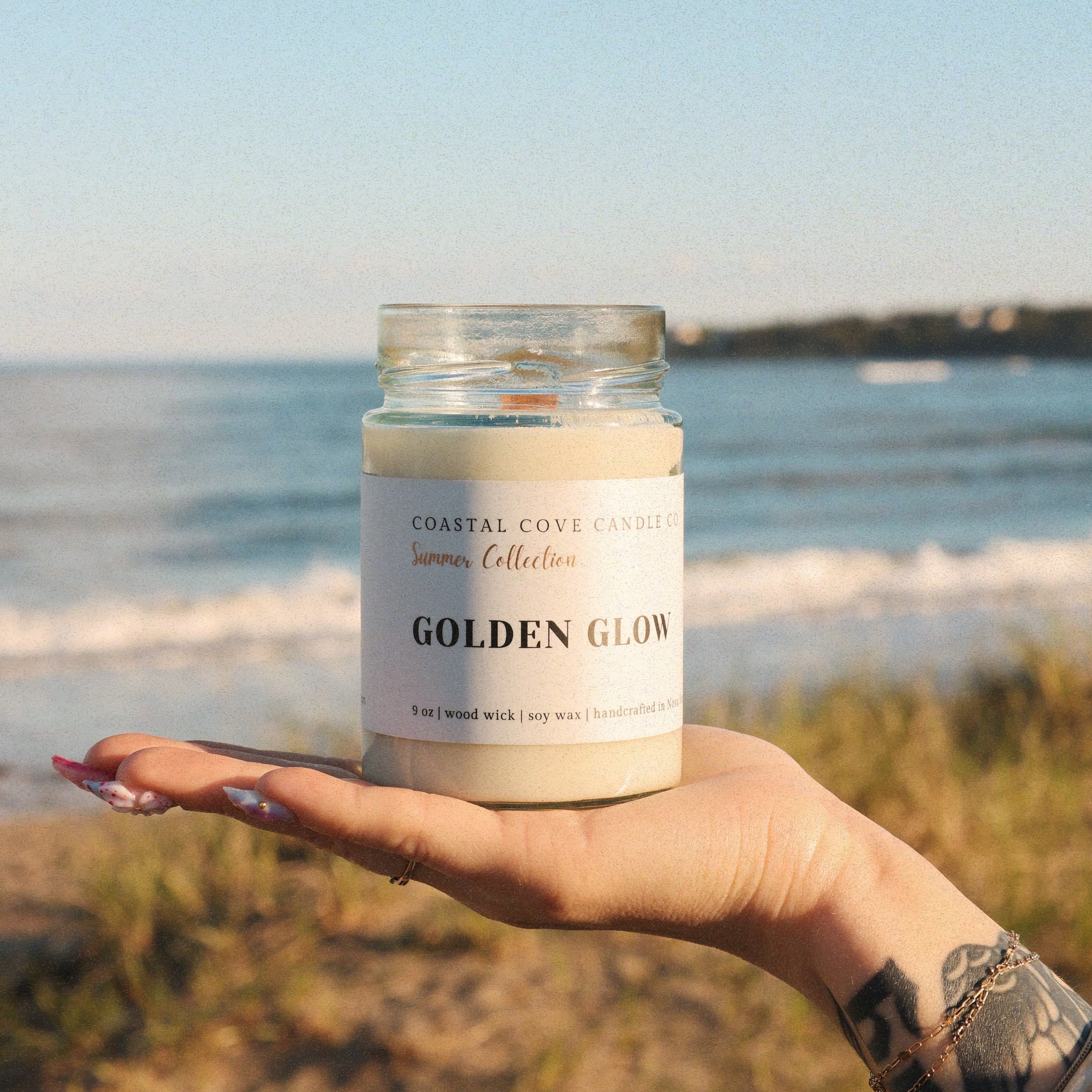 Hand holding a candle labeled 'Golden Glow' with a beach background