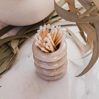 A small stacked ceramic holder in a speckled beige finish, filled with wooden matchsticks with white tips, styled on a light marble surface with dried leaves and a ceramic vase in the background.