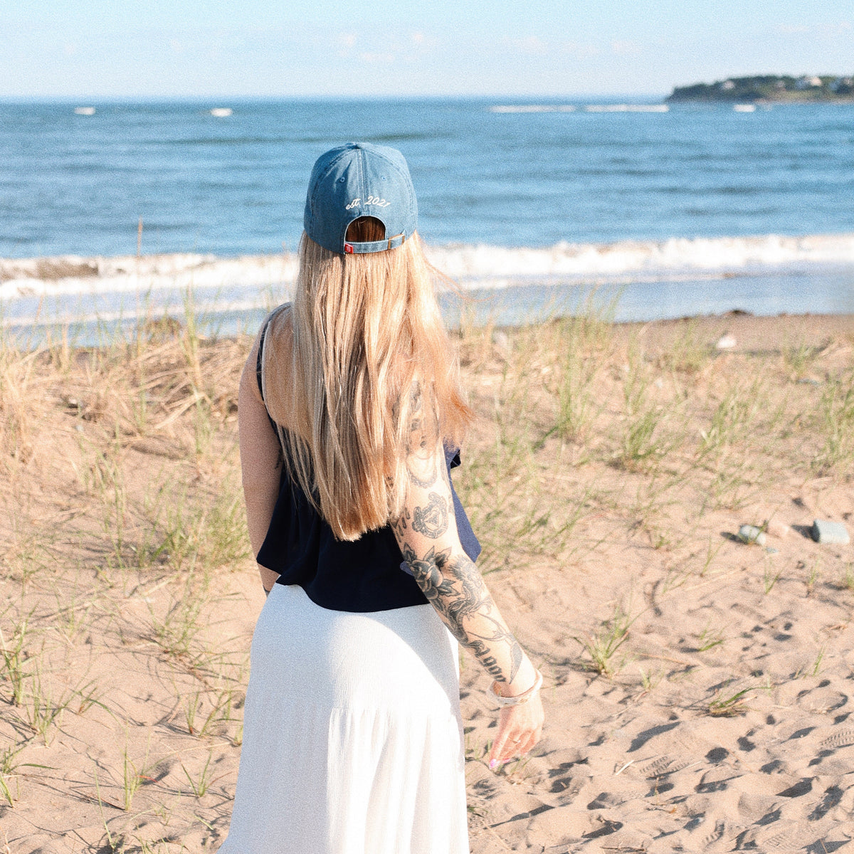 Person with tattoos wearing a black sleeveless top and white pants on a beach.