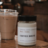A jar candle labeled 'COFFEE HOUSE' by Coastal Cove Candle Co sits on a rustic wooden table next to an iced coffee in a blurred cafe setting.