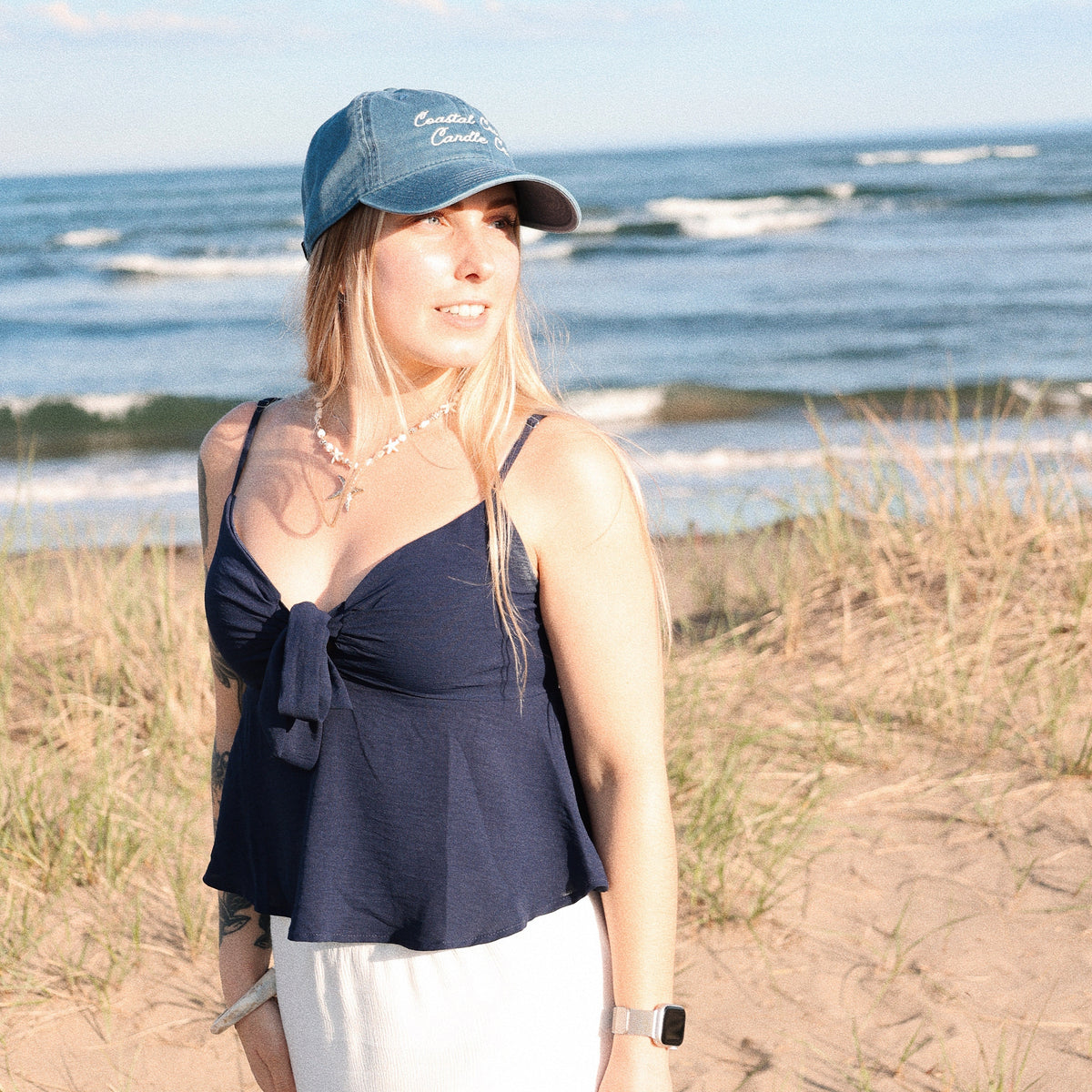 Woman standing on a sandy beach wearing a blue cap and navy top with white pants.