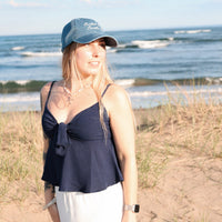 Woman standing on a sandy beach wearing a blue cap and navy top with white pants.