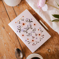 Notebook with floral design on a wooden table with coffee, spoon, and flowers.