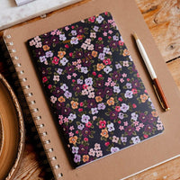 A spiral-bound notebook with a dark floral patterned cover lies on a kraft paper sleeve on a wooden table, with a pen beside it and matching stationery items partially visible at the top.
