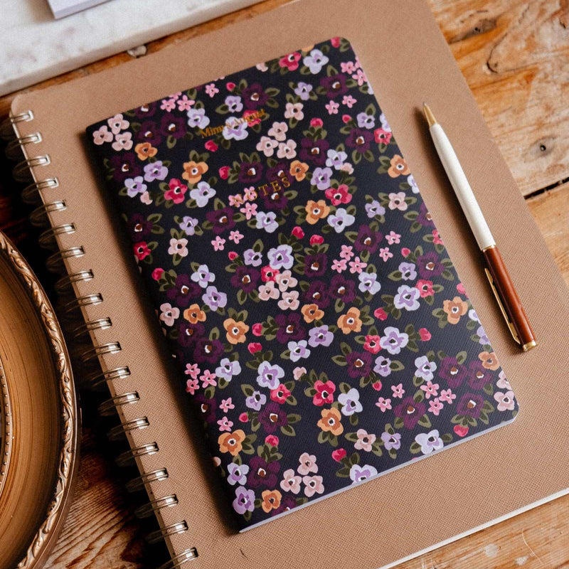 A spiral-bound notebook with a dark floral patterned cover lies on a kraft paper sleeve on a wooden table, with a pen beside it and matching stationery items partially visible at the top.