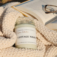 Candle labeled 'Cottage Nights' from Coastal Cove Candles on a textured surface with an open book and glasses.