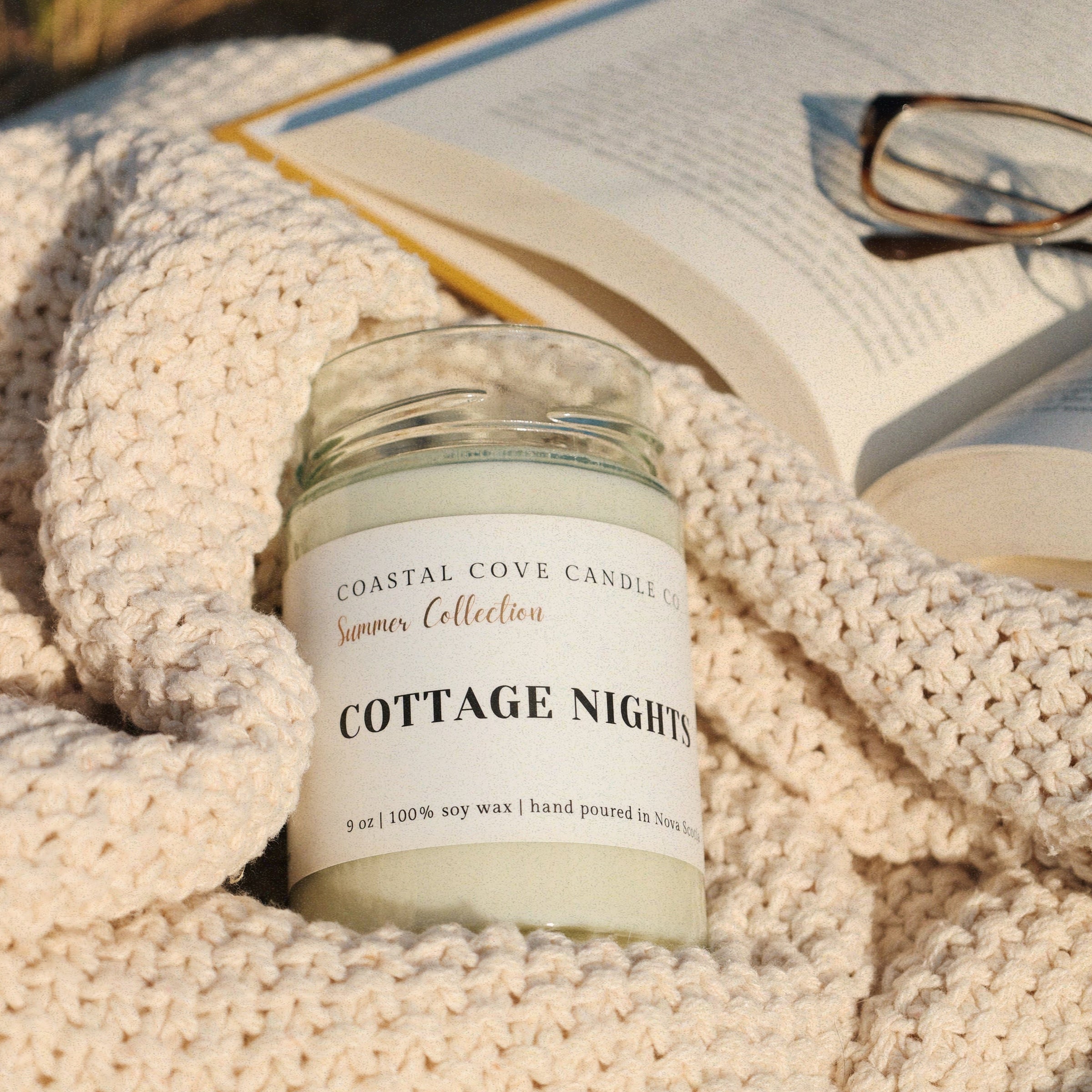 Candle labeled 'Cottage Nights' from Coastal Cove Candles on a textured surface with an open book and glasses.