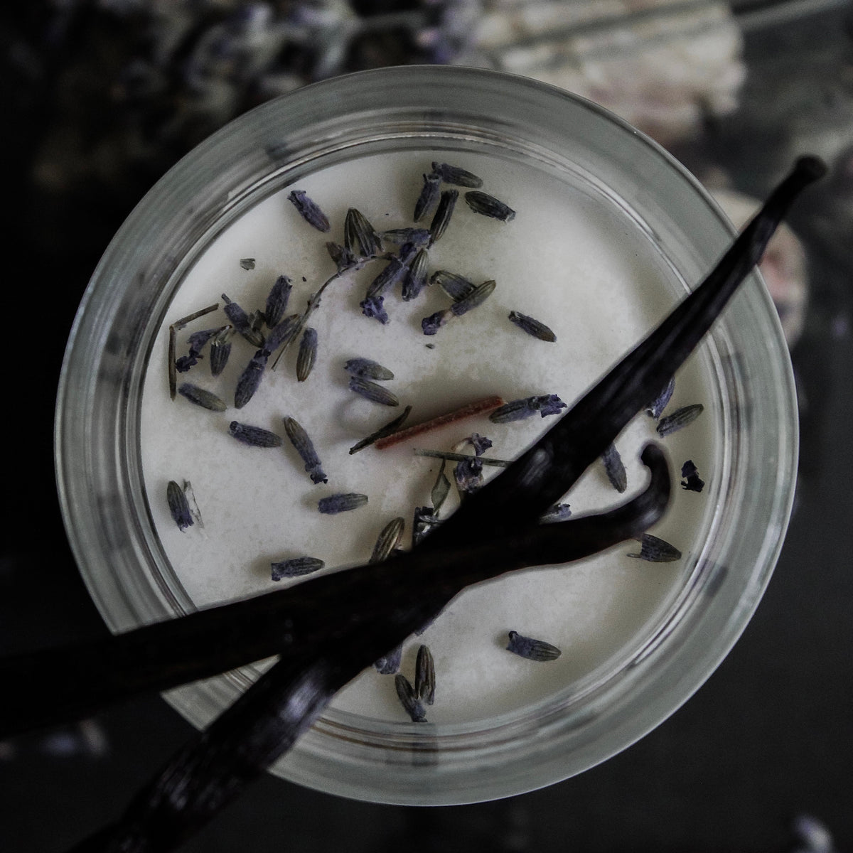A top-down view of a clear candle glass jar, topped with dried lavender buds and two dark vanilla bean pods, set against a dark background with blurred lavender sprigs.
