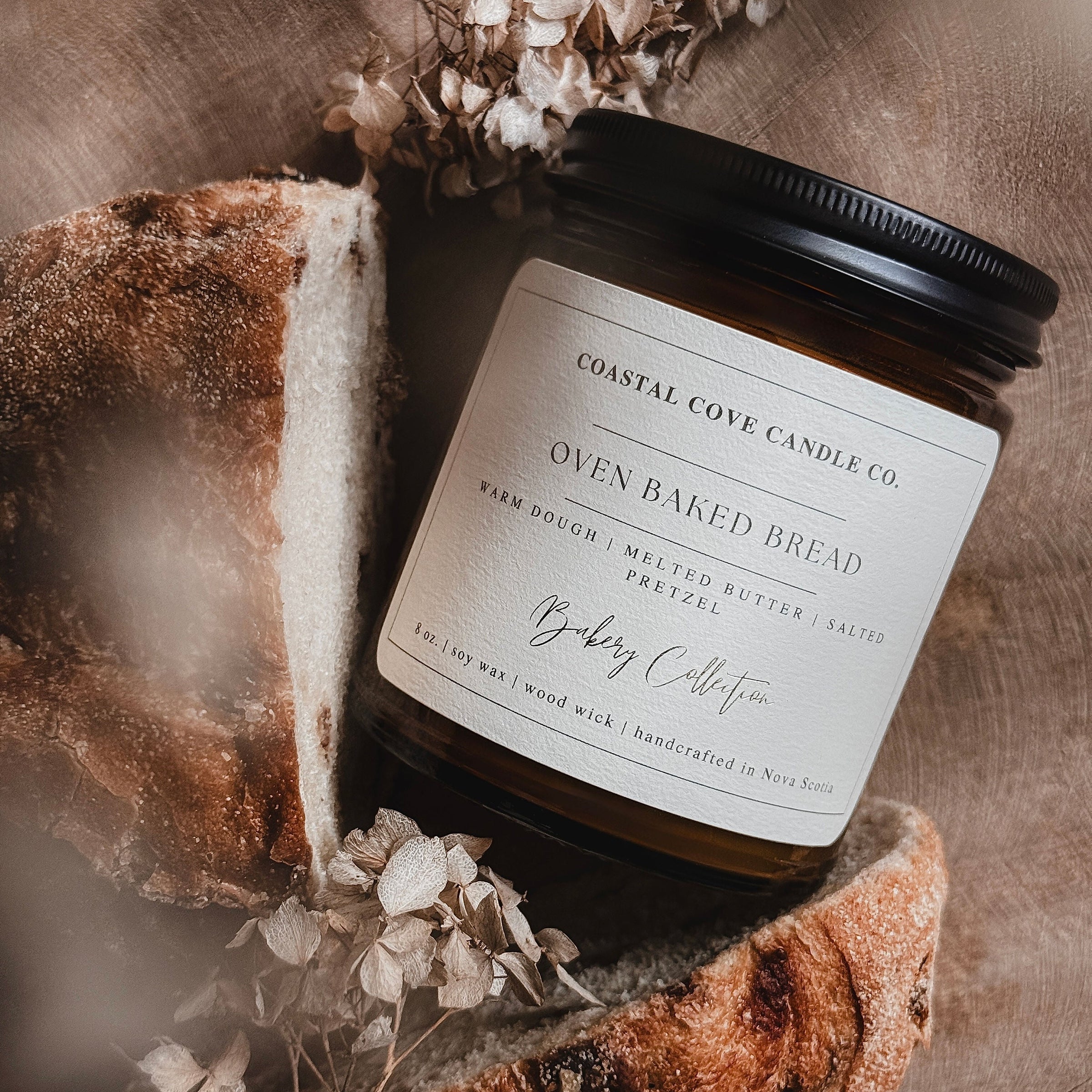 Jar of 'Oven Baked Bread' candle from Coastal Cove Candle Co on a rustic wooden surface with bread and dried flowers.