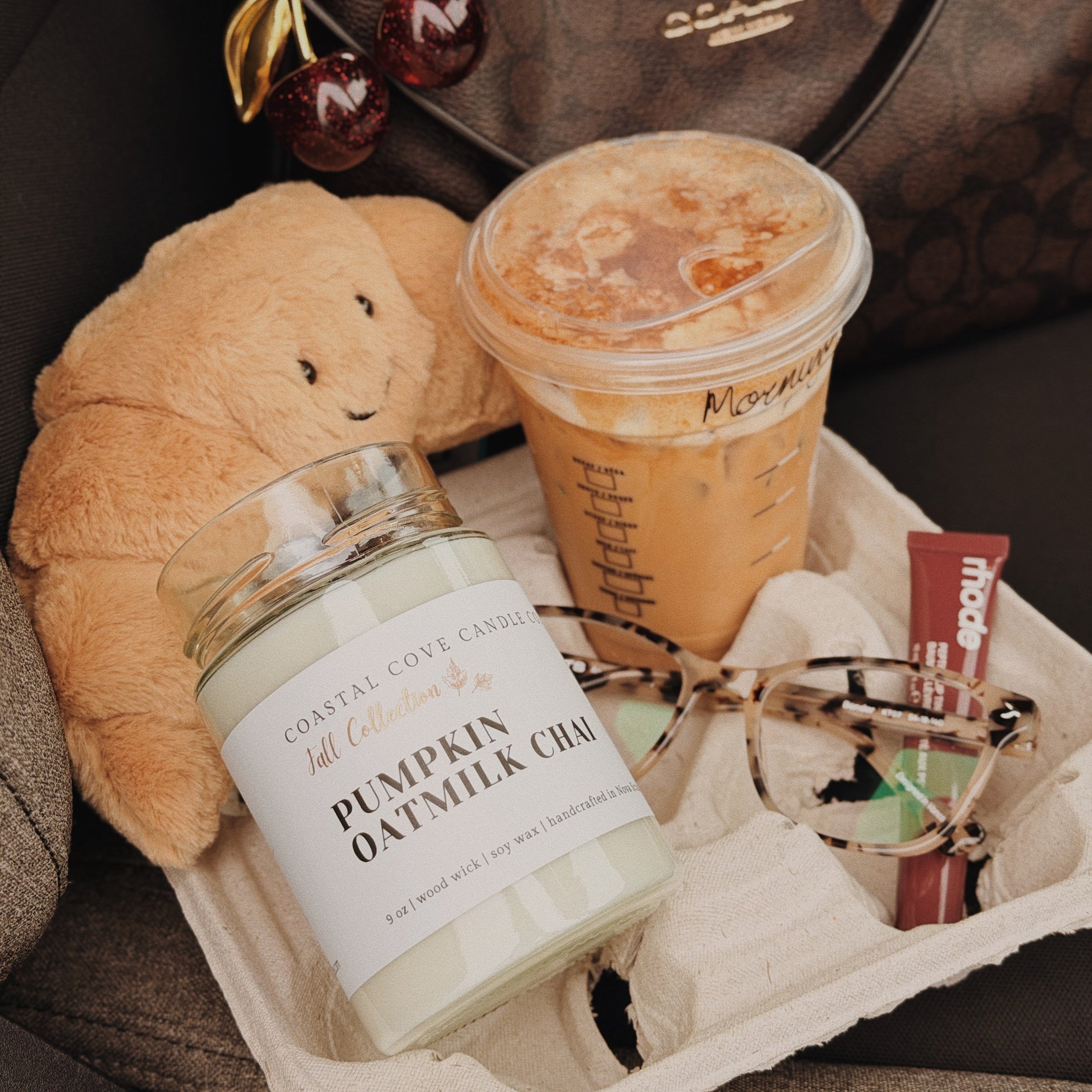 A styled tray holding a jar candle labeled 'Pumpkin Oatmeal Chai' by Coastal Cove Candle Co., an iced coffee cup with a lid, eyeglasses, and a plush cream croissant toy, set against a brown patterned bag and dark surface.