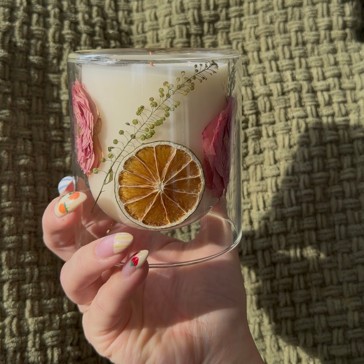 Hand holding a glass container with dried flowers and citrus slices against a textured brown background