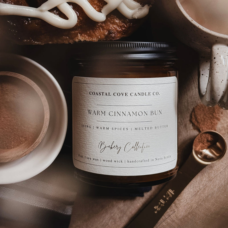 Coastal Cove Candle Co. Warm Cinnamon Bun candle with cinnamon bun and cinnamon sticks on a wooden surface