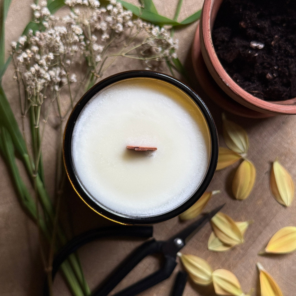 Candle in a brown container with flowers, leaves, and scissors on a brown surface