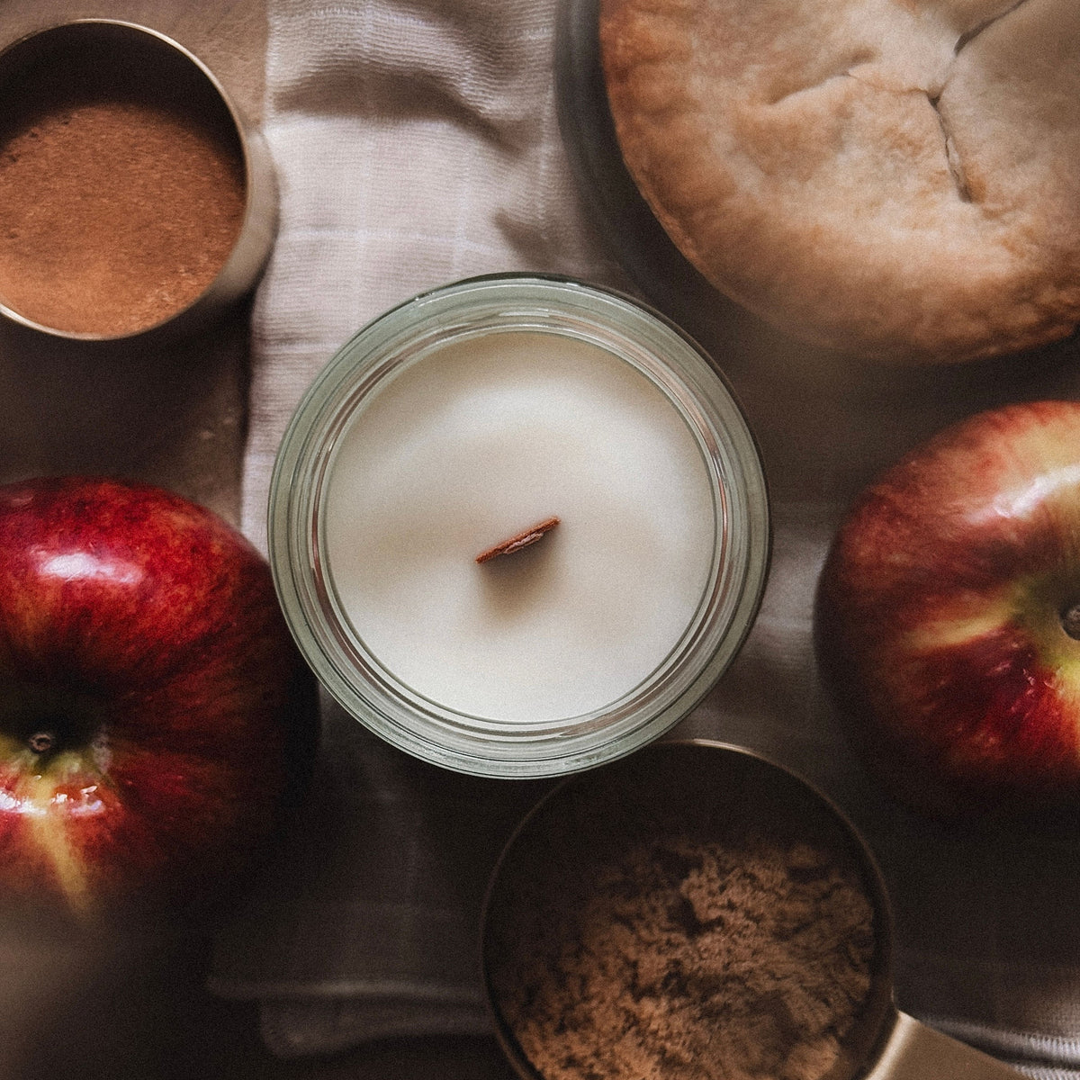 Candle in a jar surrounded by apples, a pie, and spices on a wooden surface.