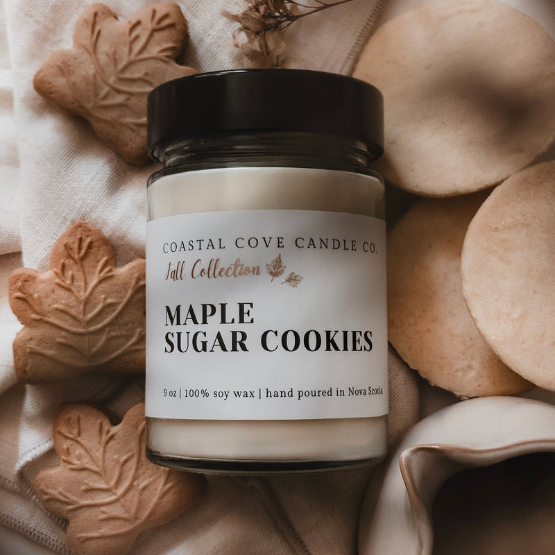 Jar of 'Maple Sugar Cookies' candle from Coastal Cove Candle Co. on a textured surface with cookies and leaves.