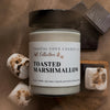 Jar of 'Toasted Marshmallow' candle from Coastal Cove Candle Co. on a wooden surface with marshmallows and chocolate.