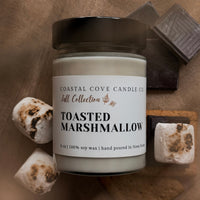 Jar of 'Toasted Marshmallow' candle from Coastal Cove Candle Co. on a wooden surface with marshmallows and chocolate.
