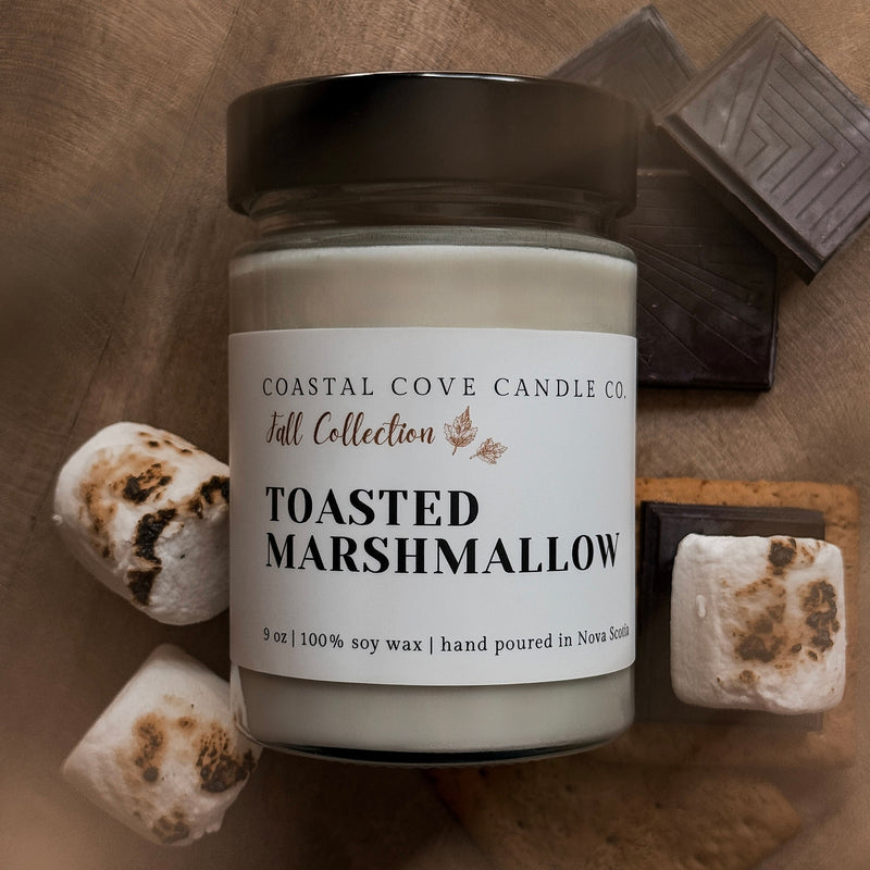 Jar of 'Toasted Marshmallow' candle from Coastal Cove Candle Co. on a wooden surface with marshmallows and chocolate.