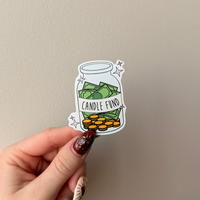 Hand holding a sticker of a 'Candle Fund' jar against a plain background