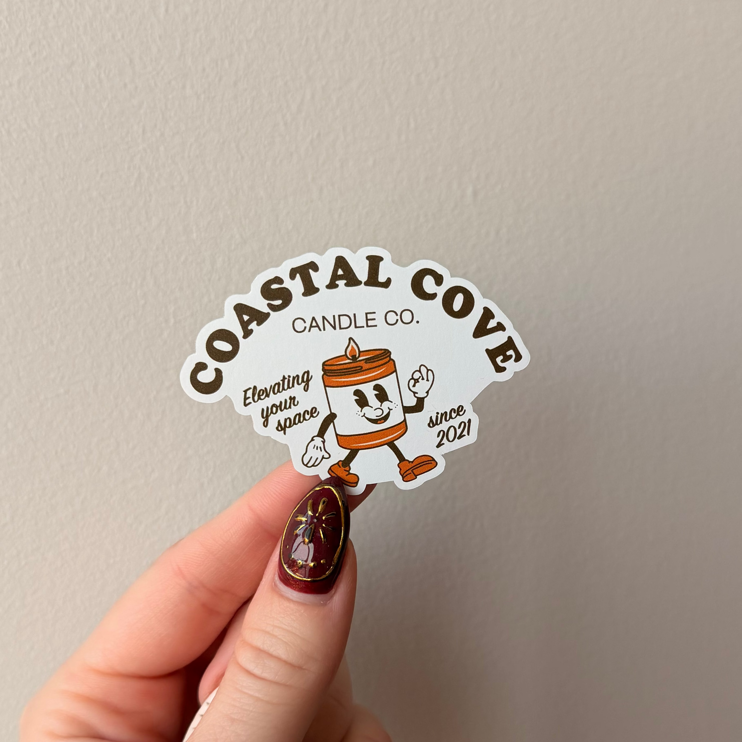 Hand holding a sticker with 'Coastal Cove Candle Co.' design against a plain background
