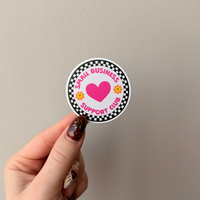 Hand holding a circular sticker with a heart and text on a plain background