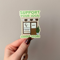 Hand holding a sticker with 'Support Your Local Candle Store' text against a plain background