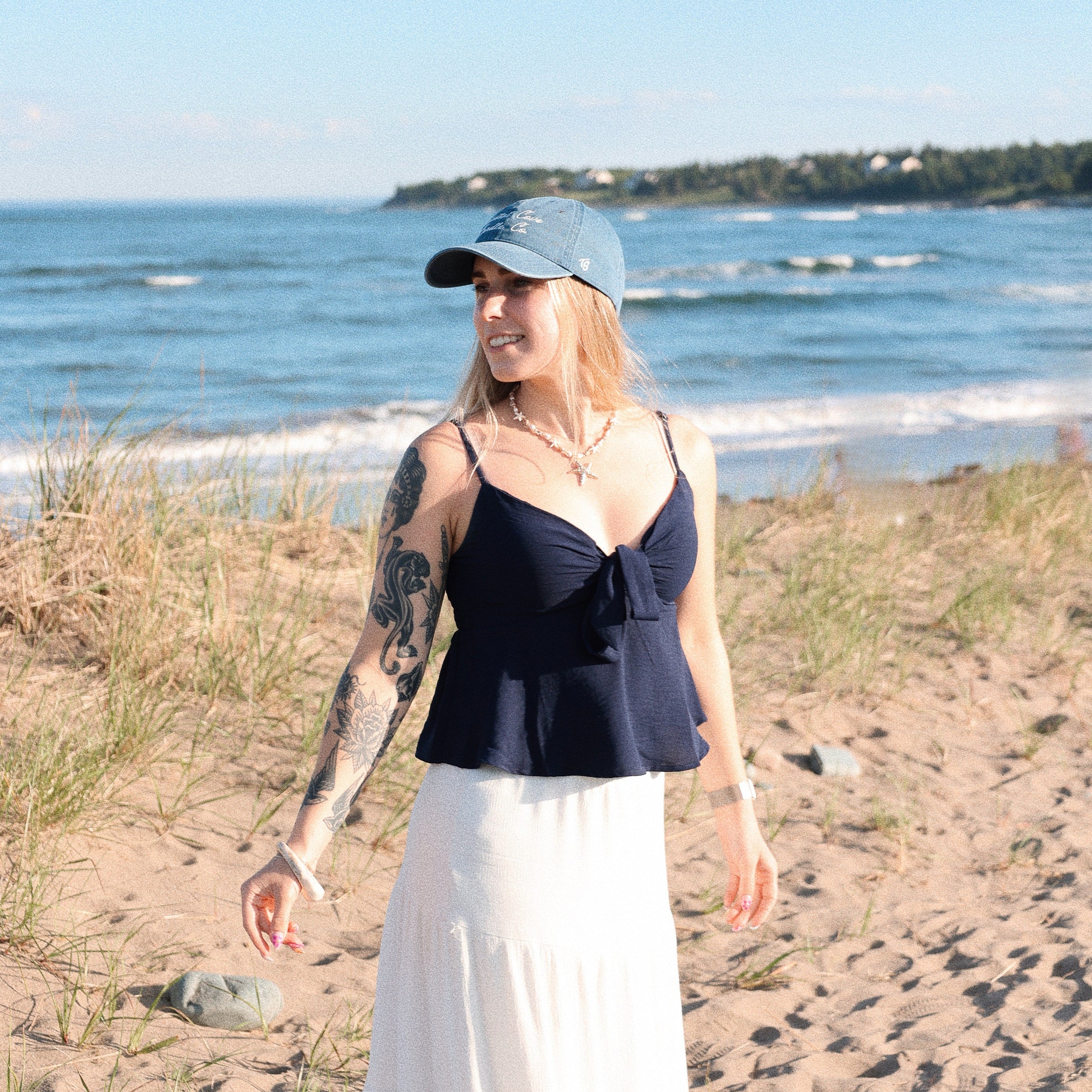 Woman standing on a beach wearing a black top and white skirt with a blue cap.