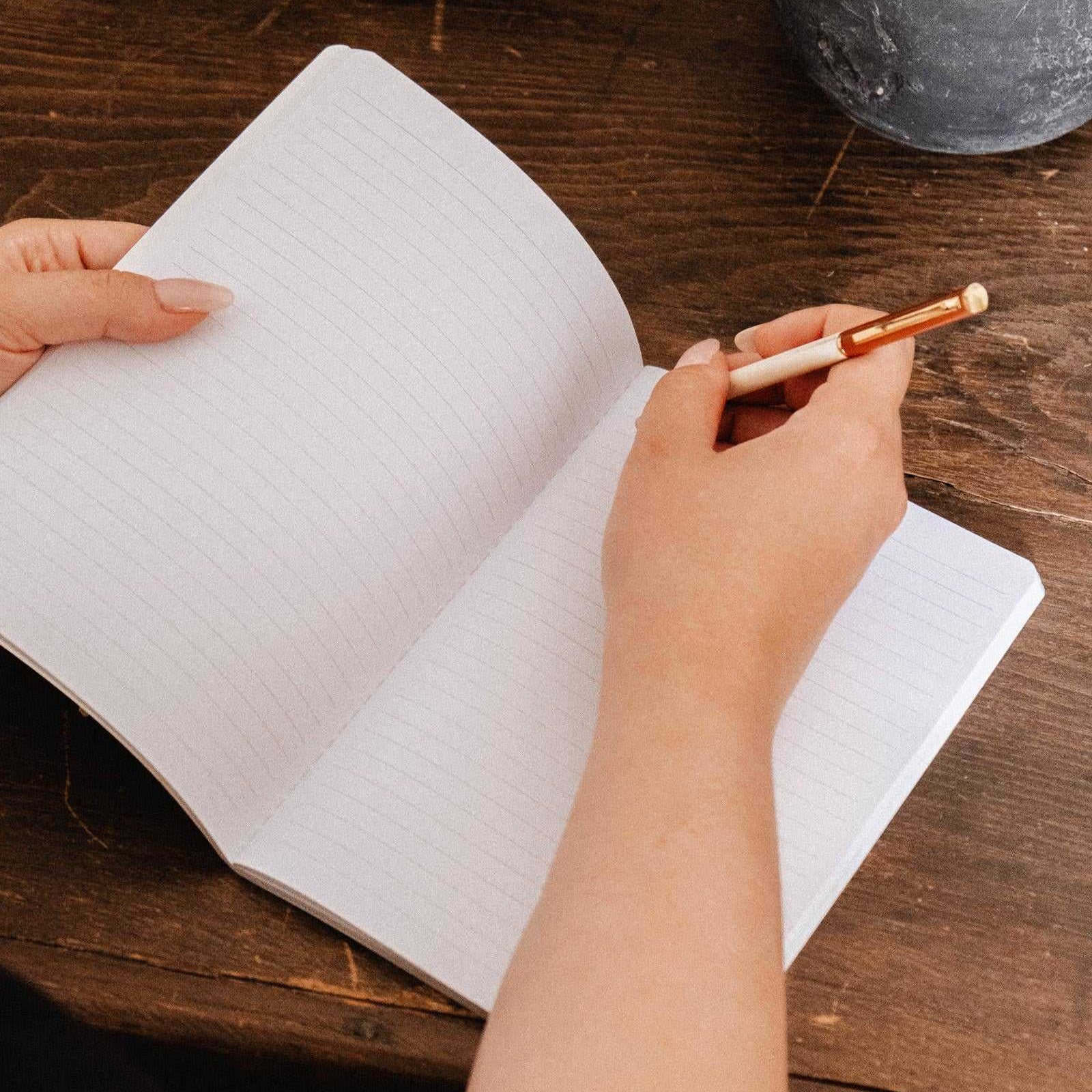 A top-down view of a person's hands writing in an open lined notebook with a white and rose gold pen, set on a dark wooden table with decorative props.