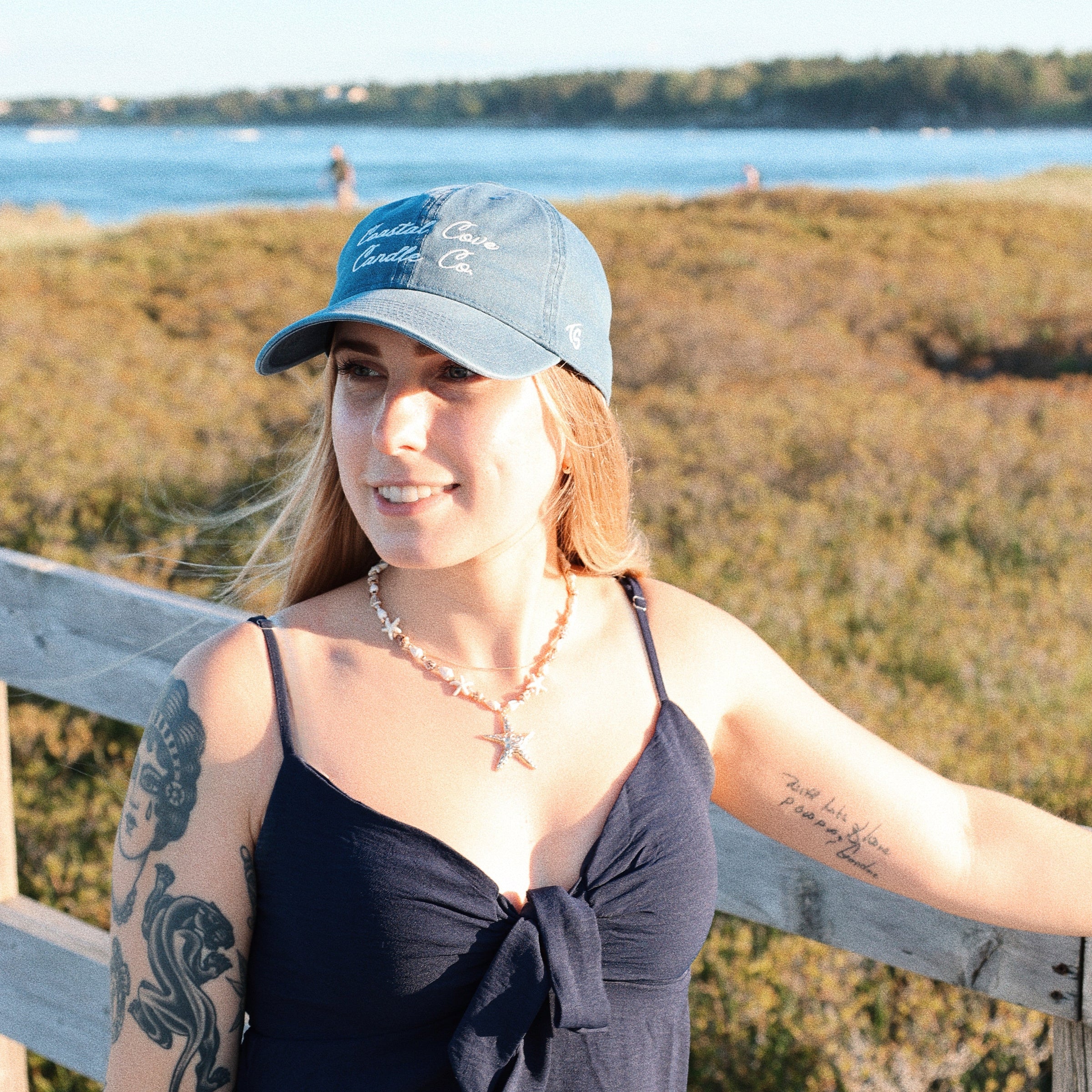 Woman wearing a blue cap and black tank top standing by a wooden fence with a scenic background.