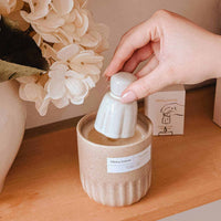 Hand placing a white ceramic object into a beige candle on a wooden surface with flowers in the background.