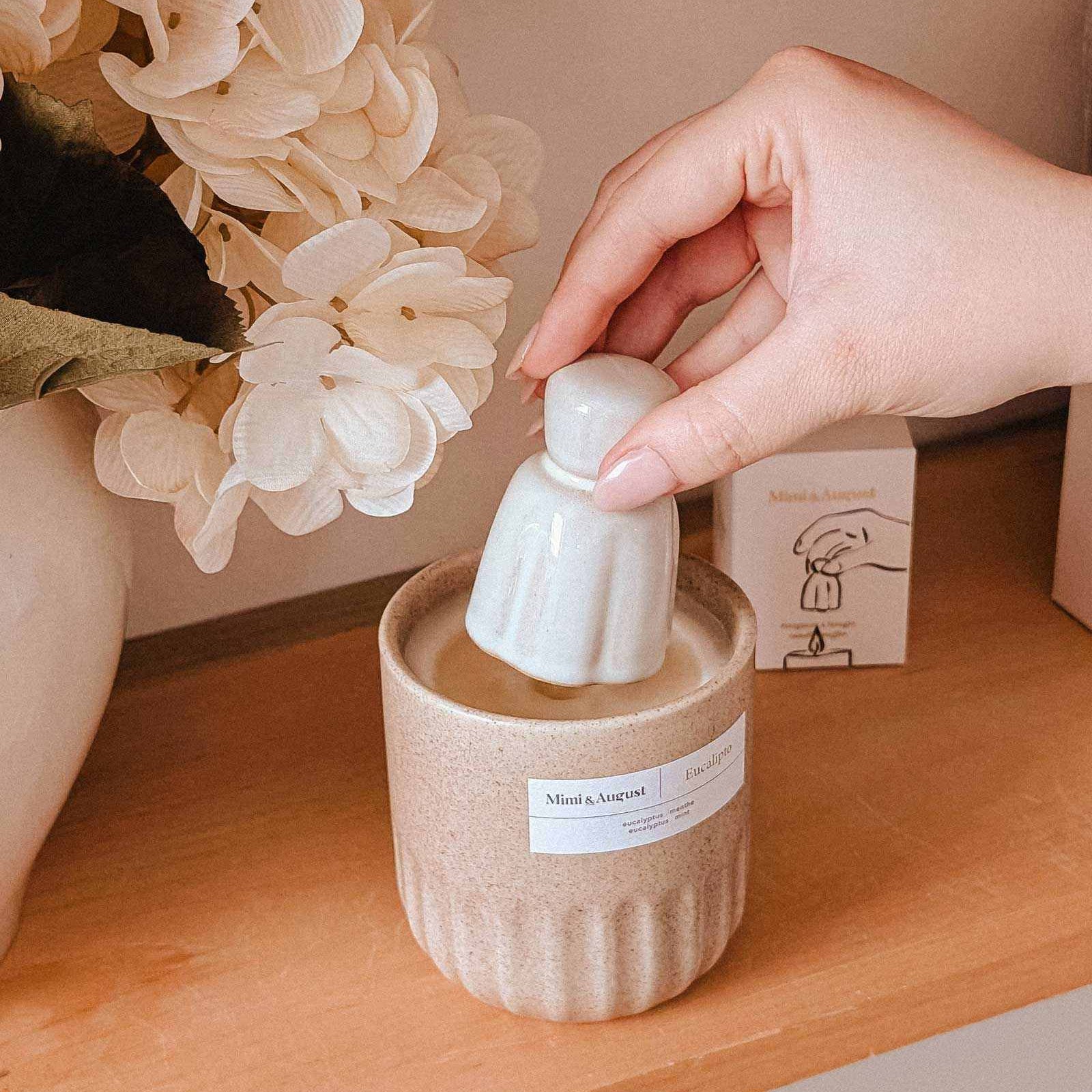 Hand placing a white ceramic object into a beige candle on a wooden surface with flowers in the background.