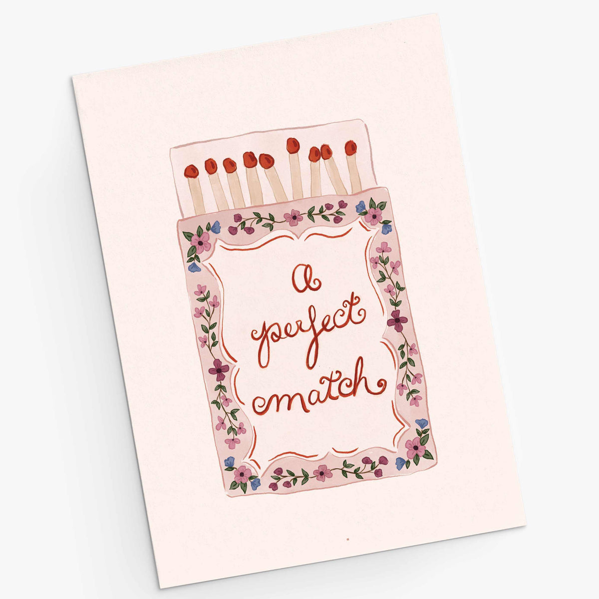 A square greeting card featuring an illustration of a matchbox with the phrase 'a perfect match' in cursive. The card is light pink with a floral border and red match heads, photographed on a clean white background.