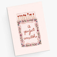 A square greeting card featuring an illustration of a matchbox with the phrase 'a perfect match' in cursive. The card is light pink with a floral border and red match heads, photographed on a clean white background.