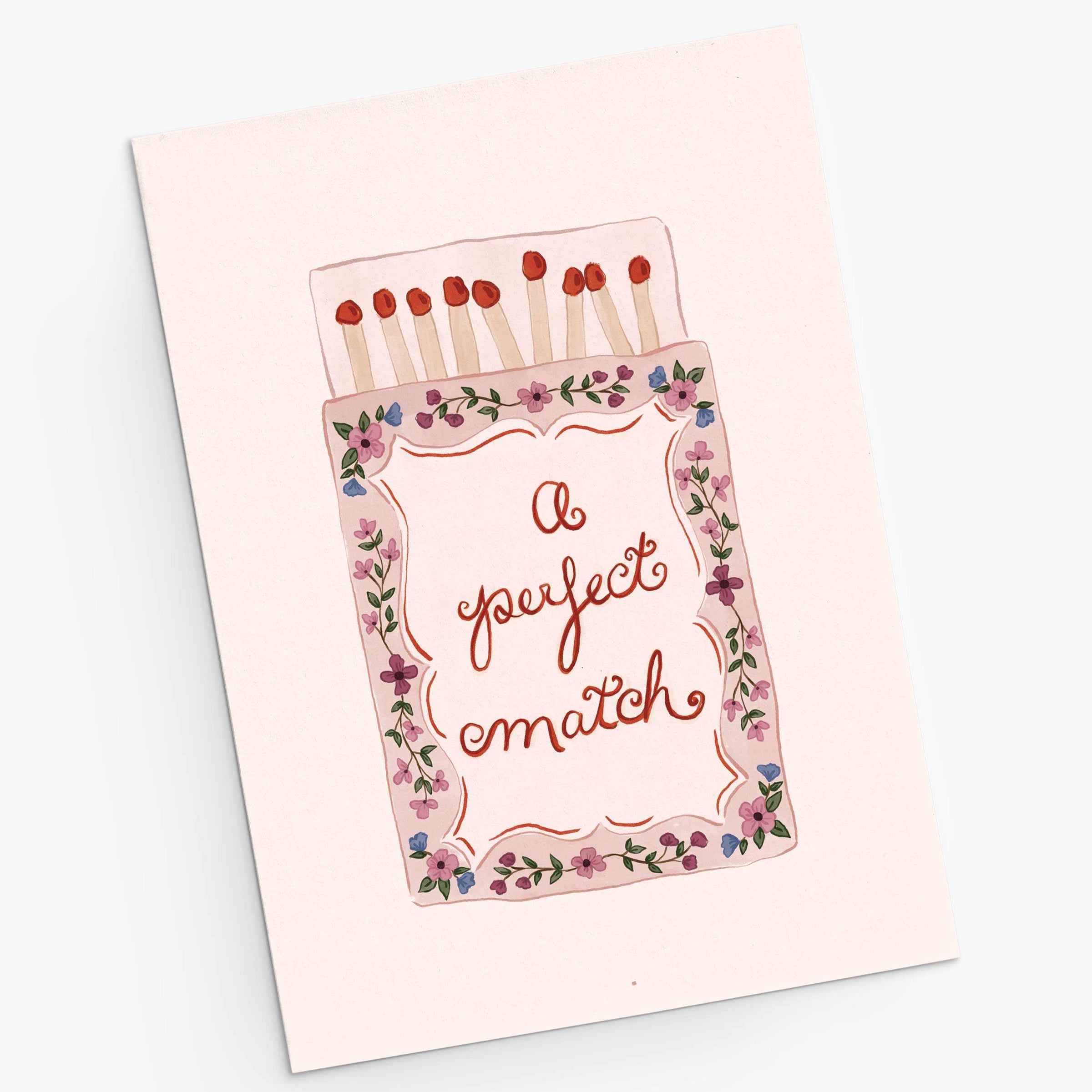 A square greeting card featuring an illustration of a matchbox with the phrase 'a perfect match' in cursive. The card is light pink with a floral border and red match heads, photographed on a clean white background.
