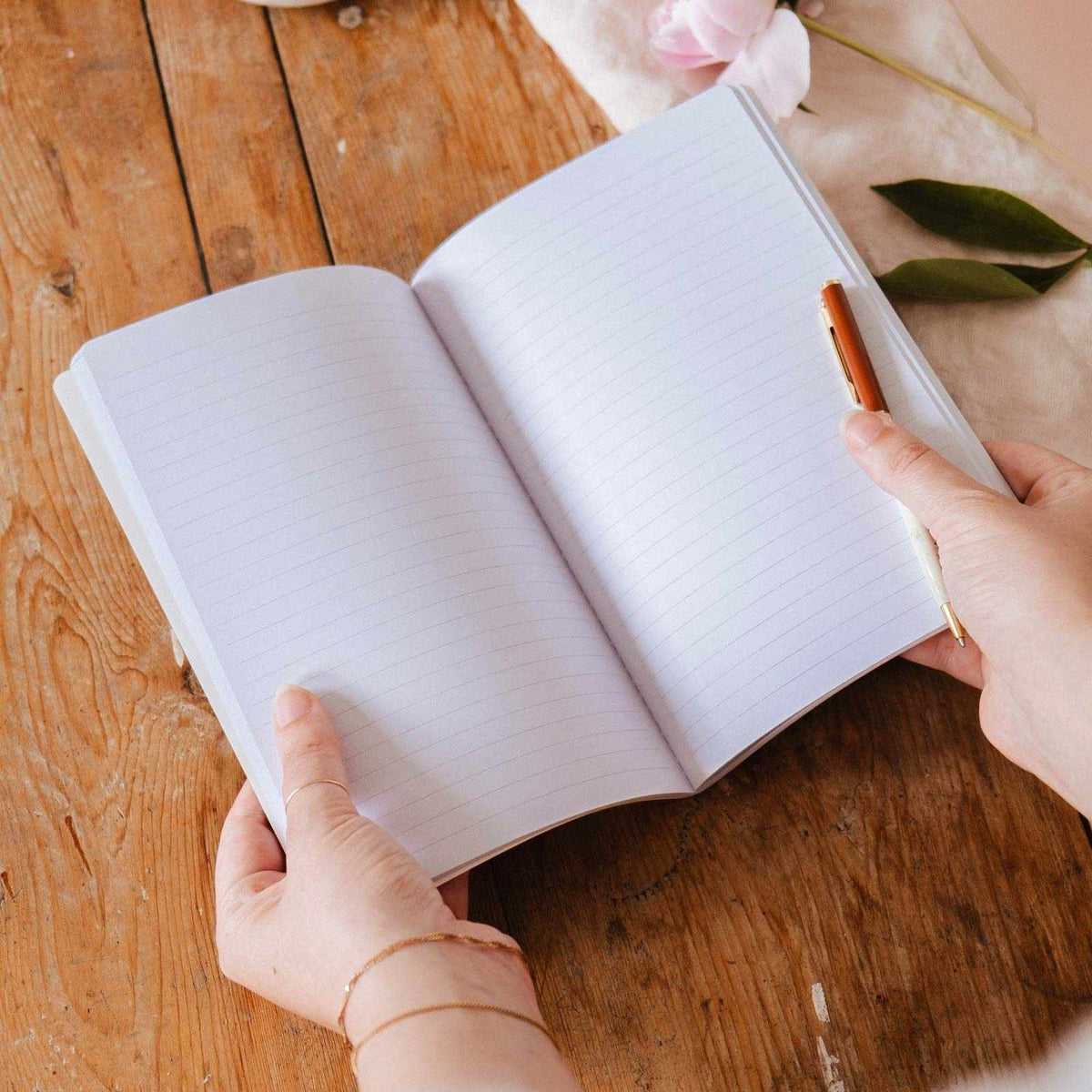 Person holding an open notebook with a pen on a wooden surface, with a cup and flowers in the background.