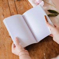 Person holding an open notebook with a pen on a wooden surface, with a cup and flowers in the background.