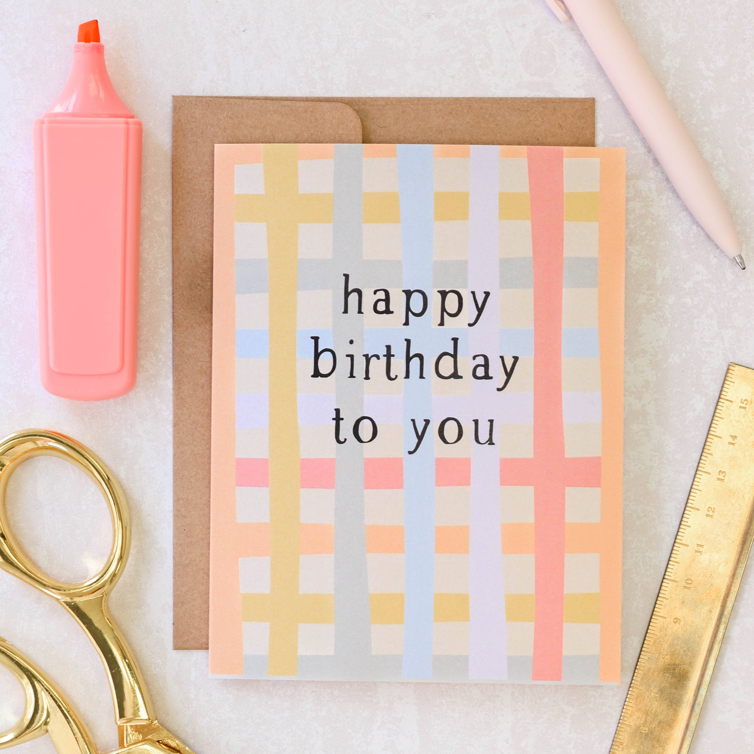 Birthday card with pastel checkered pattern and 'happy birthday to you' text, surrounded by stationery items on a light background.